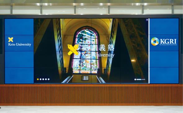 写真：55V型・縦４面、横６面の24面の大型マルチスクリーンシステムの左右縦３面は青背景、左に「Keio university」、右に「KGRI」の文字が映し出されている。中央16面には教会のステンドグラスの背景、慶応義塾大学のロゴとともに「慶應義塾」という文字が映し出されている。