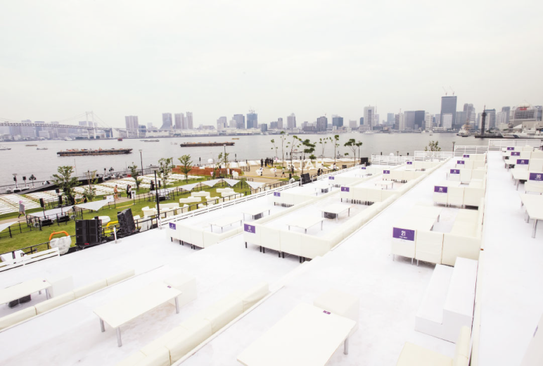 写真:東京湾前の会場。遠くにビル群があり、手前に海、芝生の陸、段差をつけた観客席には白いシートと机が置かれている広いVIPエリアがある。