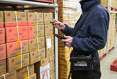 写真:入荷検品している作業員の様子。手に持ったスキャナで箱のバーコードを読み取り首から掛けたタフパッドで商品情報を確認し、腰元にベルトで巻いたプリンタからシールを出す。