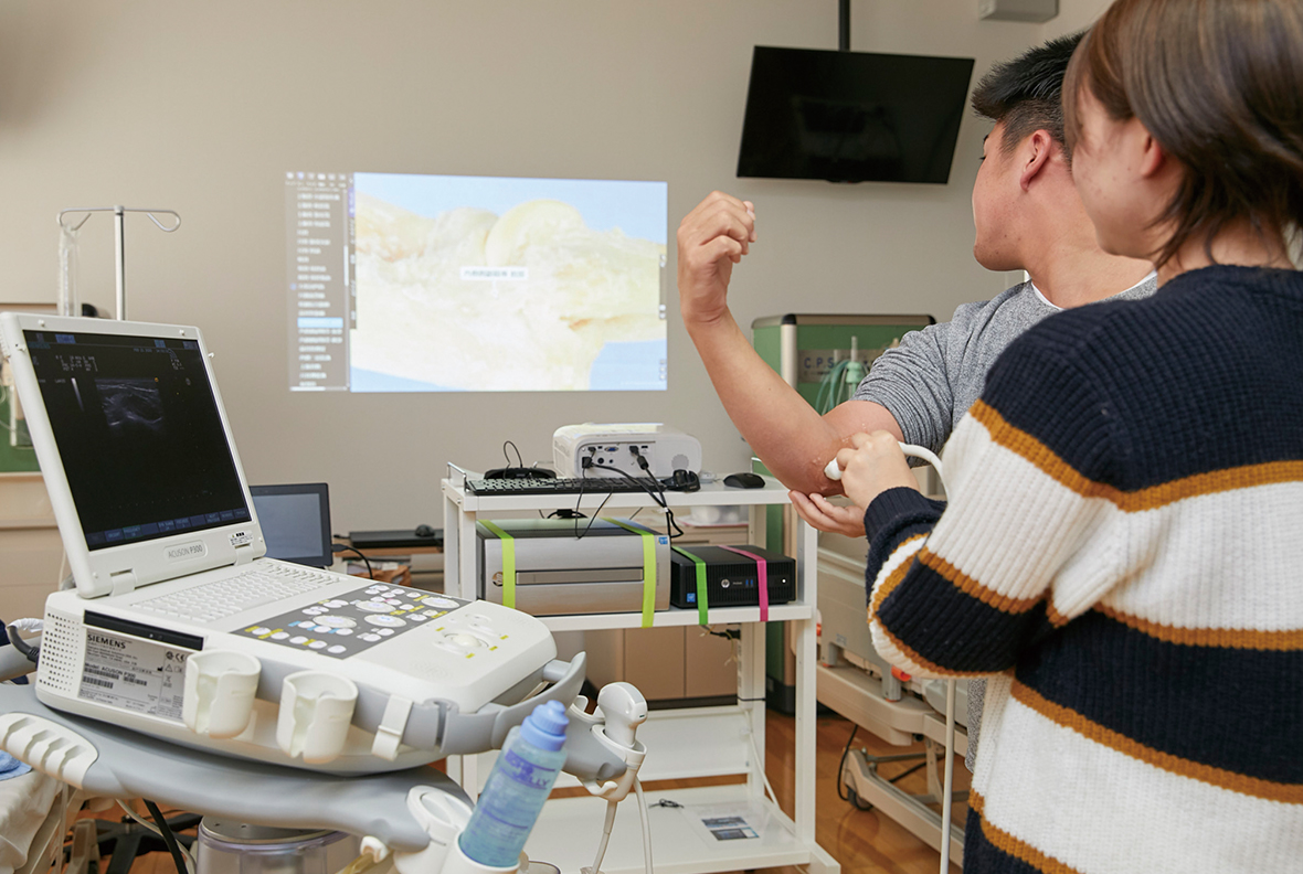写真:女性が男性の肘の内側に超音波エコー機を当て、壁に映し出された解剖映像を確認している様子。
