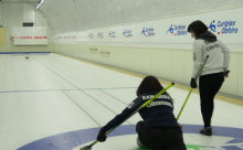 写真：二人の女性カーリング選手が競技場でカーリングをする様子。左の選手がストーンを持ち投げようとしており、右の選手はその右横に立ちストーンを見ている。